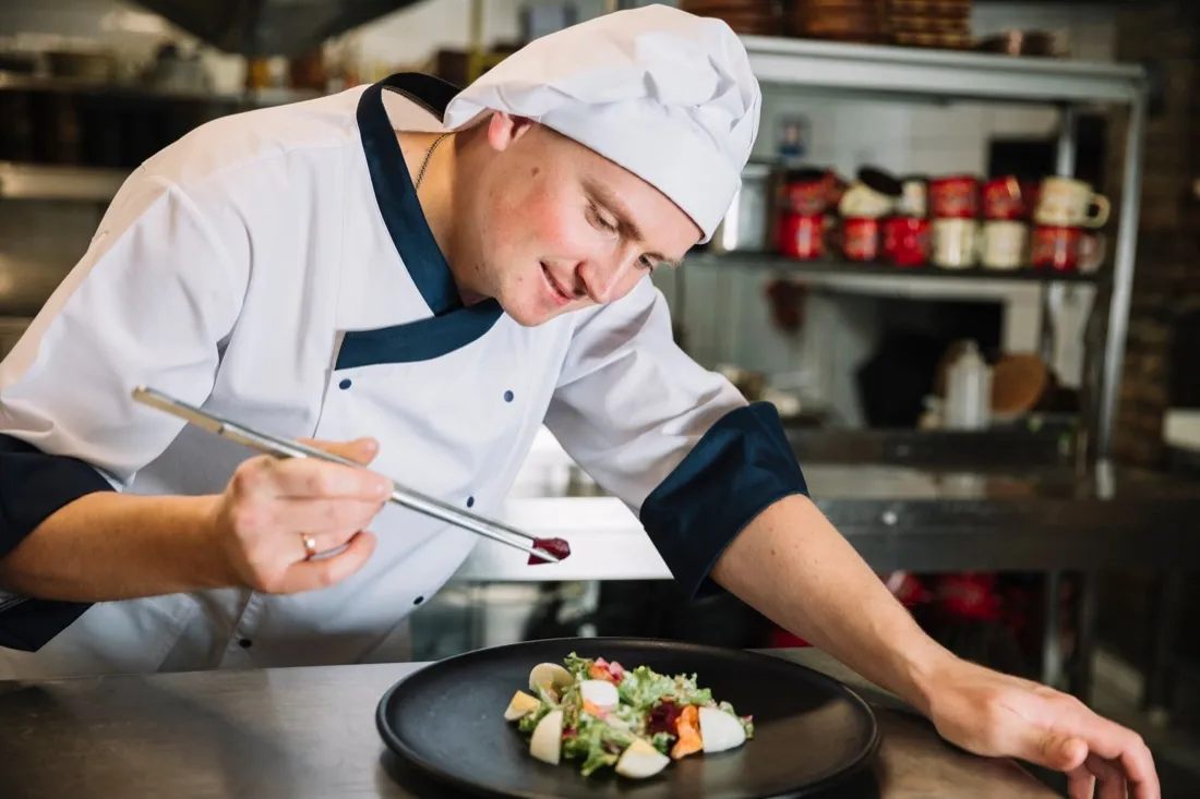 A chef leaning over a dark bowl, finishing a plate with tweezers