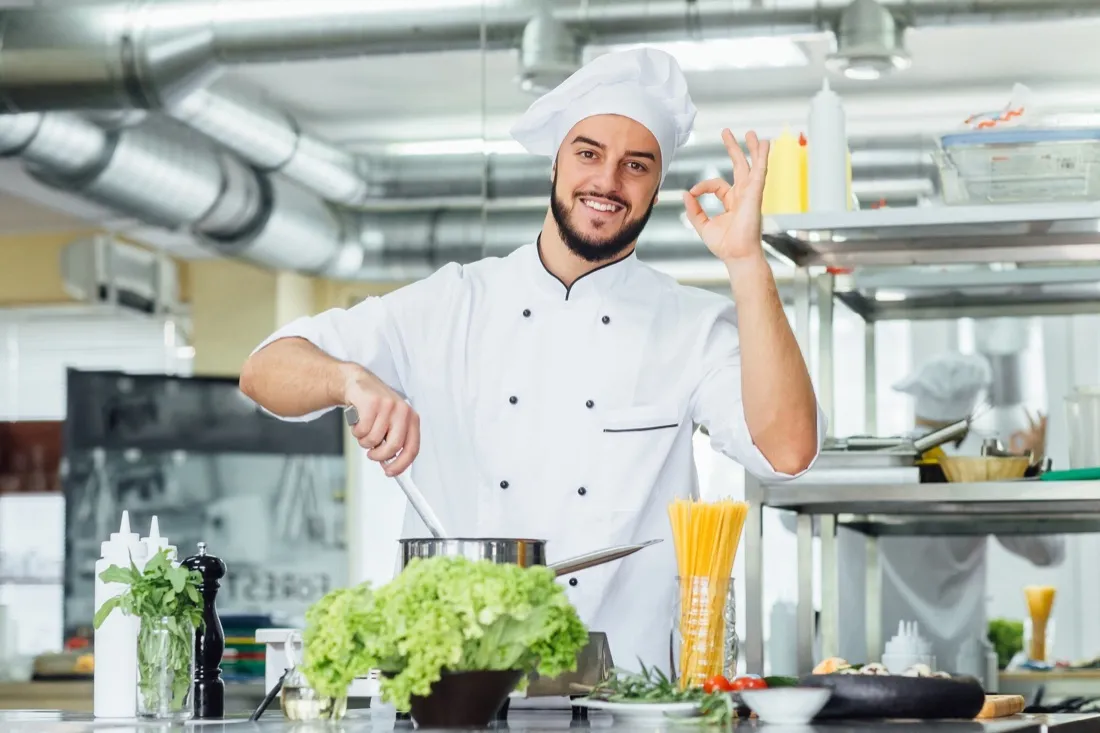A smiling chef stirring a pot and holding up an OK sign